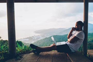 Thoughtful young man looking away on beautiful nature landscape while sitting with laptop computer outdoors. Blogger guy catch inspiration  writing article on netbook during vacation holidays in Thai