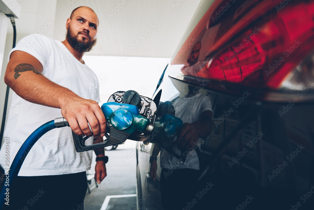 Fototapeta premium Serious bearded hipster guy filling talk of automobile for driving holding hose and nozzle on service station,young male owner of car refill vehicle with petroleum, climate pollution concept.