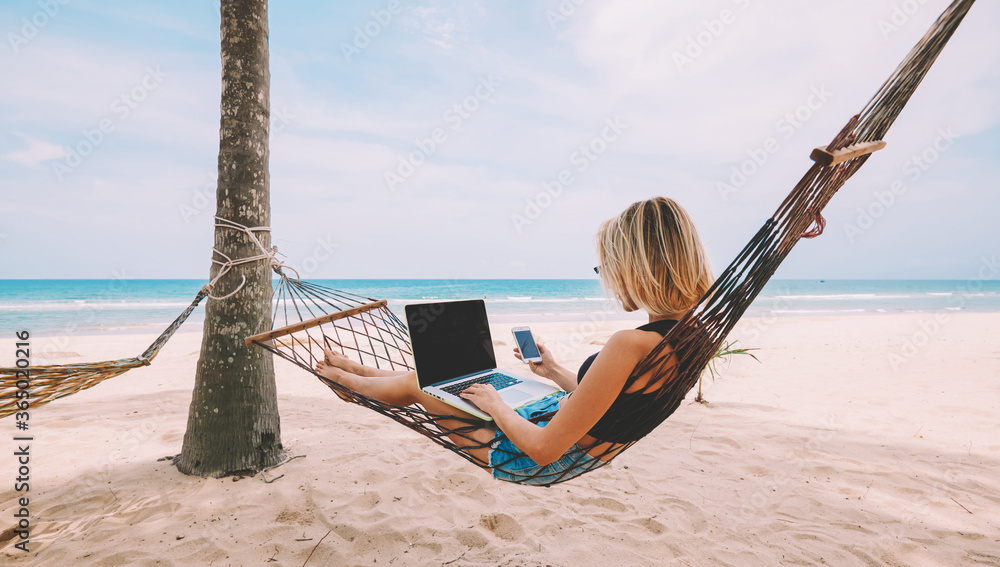Young woman tourist reading notification on smartphone in roaming while lying on hammock and working remote at laptop computer with mock up area for your content during summer vacation on ocean beach - obrazy, fototapety, plakaty 