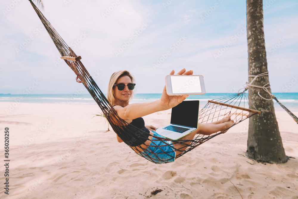 Cheerful blonde female freelancer showing smartphone with blank screen for your internet web page while working at netbook on hammock during vacations distance job on tropical sea shore in summer time