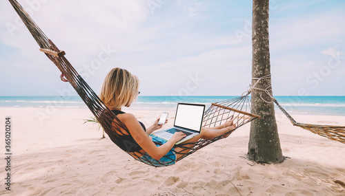 Female freelancer entrepreneur lying on hammock and doing remote business work at modern laptop computer with blank screen area for web page on smartphone during summer vacation on tropical seashore