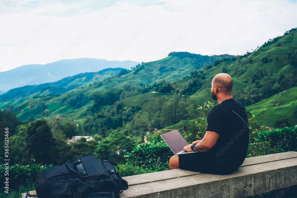 Young man freelancer sitting on hill with modern laptop computer for remote work while enjoying amazing view of high mountains with green plantation and vegetation during summer trip