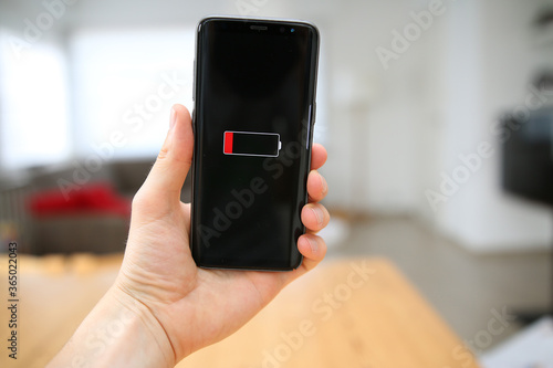 A single red bar in a battery icon on the touch screen of a mobile phone, held up by a male hand. A warning about the empty or low level of the battery power in the device. A charging concept.