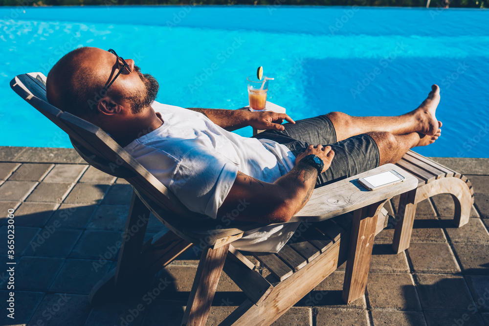 Young bearded man in sunglasses sunbathing on sunbed with cocktail near blue swimming pool enjoying hot tour during summer vacation.Businessman with tanned skin relaxing and spending time on resort - obrazy, fototapety, plakaty 