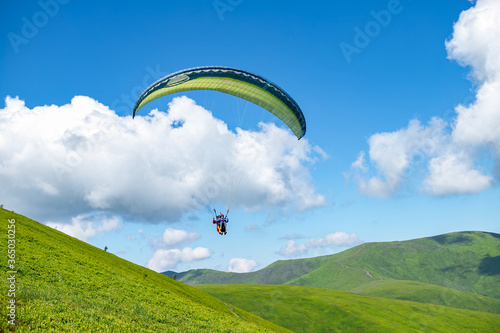 High view mountain panorama in the Carpathians. Two people on a paraglider. Paraglider fly over a mountain valley on a sunny summer day. Beautiful landscape with greenery and clouds.