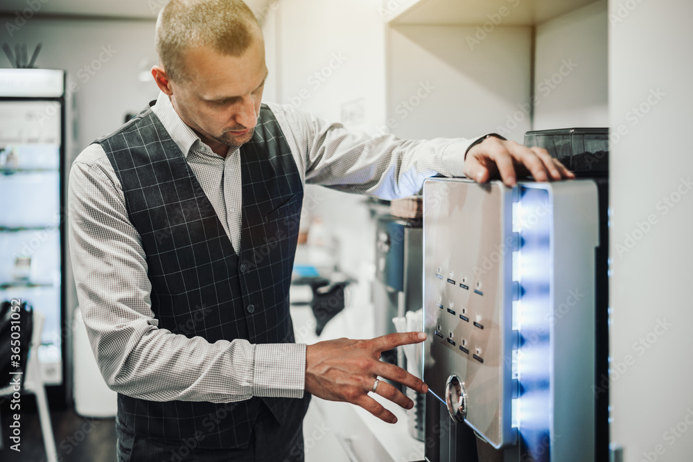 A businessman in a plaid vest is choosing which type of coffee to make and he is about to push one of the buttons of a modern automatic coffee maker machine indoors of a small bright office kitchen