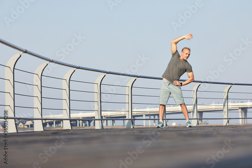 Athletic mature man in sportswear bending side with raised arm while doing warm up on embankment