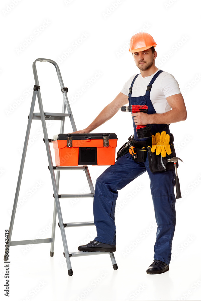 caucasian repairman worker in uniform and helmet holding cordless screwdriver and tool box near stepladder. handyman wearing tool belt isolated on studio background caucasian repairman worker in uniform and helmet holding cordless screwdriver and tool box near stepladder. handyman wearing tool belt isolated on studio background