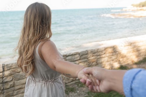An young italian woman whose face is seen with long blond hair by the sea pulls to herself a man whose arm is seen with a blue shirt
