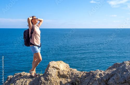 brunette woman in t-shirt with backpack stands back on rocky seaside arms over head, and looks out at the sea. Solo summer outdoor activities in fresh air. Concept of Hiking and trekking. Copy space © Tani_Bel