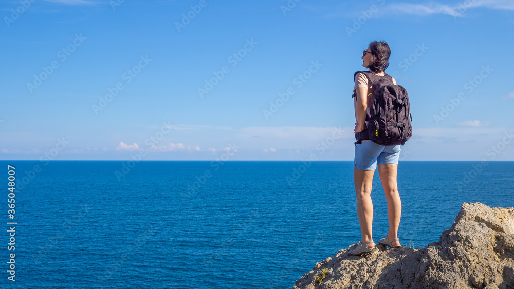 brunette woman in t-shirt with backpack stands back on rocky seaside , and looks out at the sea. Solo summer outdoor activities in fresh air. Concept of Hiking and trekking. Copy space
