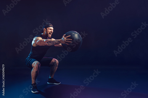 Aggressive bearded muscular sportsman is working out with a medicine ball isolated on dark studio background.