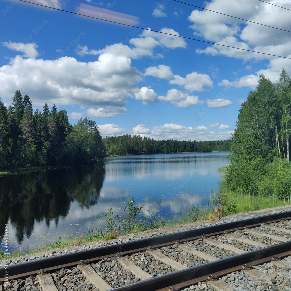 railway in the forest in Finland, lake views Finnish summer scenery  with a forest and blue sky reflection in the water, with white clouds next to railroad, from the train.