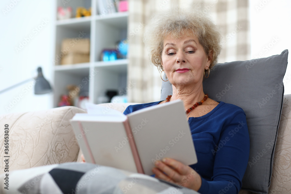 Portrait of well-educated senior female reading interesting story book. Attractive woman sitting on comfort couch at home and relaxing. Cozy interior. Hobby and spare time concept