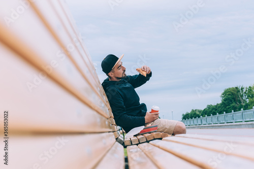 Hipster guy was sitting on a bench and having dinner with toast with coffee in a glass. Fast food in the fresh air.