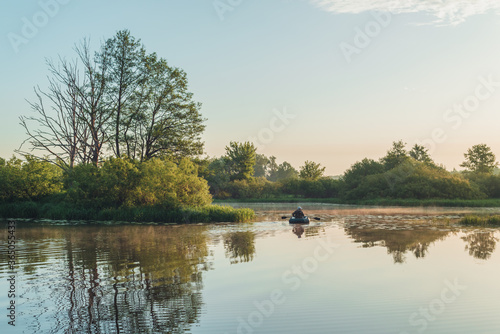 Summer landscape in Belarus. Colorful sky and reflections in the river at dawn. Man, a fisherman on a rubber boat in the middle of the river.