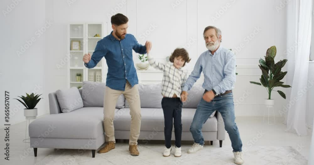 Caucasian males generations. Little cute kid with old grandfather and father dancing and moving funny in living room. Senior man with son and grandson holding hand in hand dance and listening to music Caucasian males generations. Little cute kid with old grandfather and father dancing and moving funny in living room. Senior man with son and grandson holding hand in hand dance and listening to music