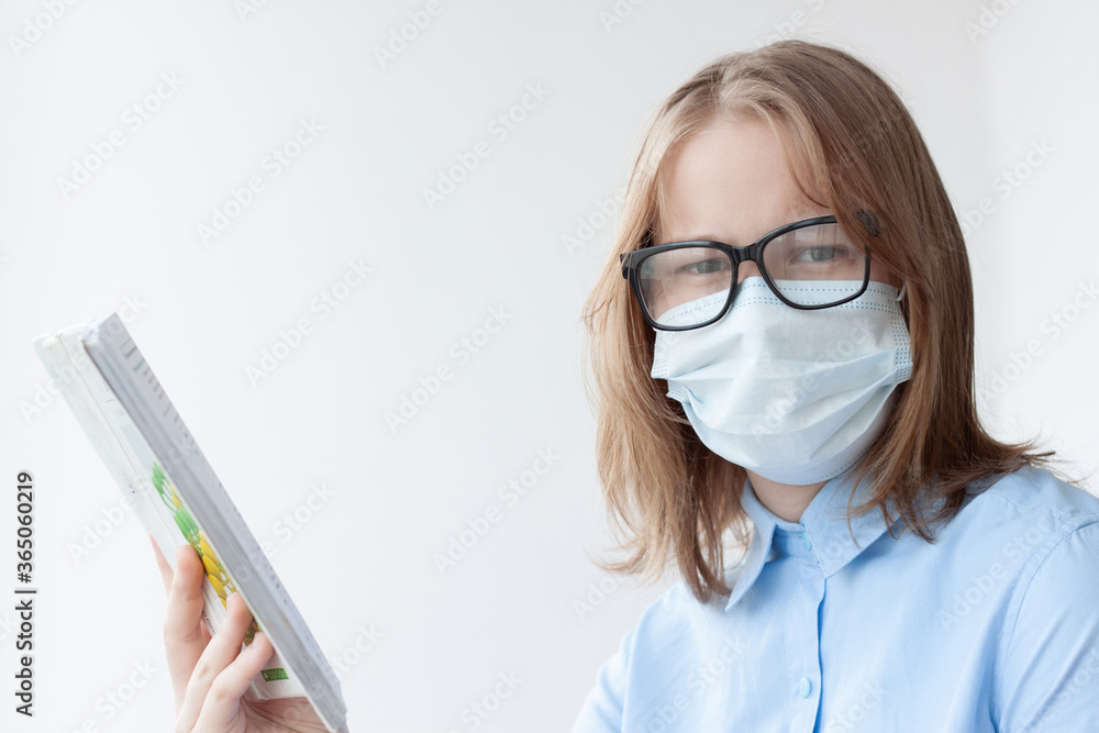 A teenage girl, blonde, in a blue shirt, a medical mask and glasses on a white background looks at the camera, holding a textbook in her hand A teenage girl, blonde, in a blue shirt, a medical mask and glasses on a white background looks at the camera, holding a textbook in her hand