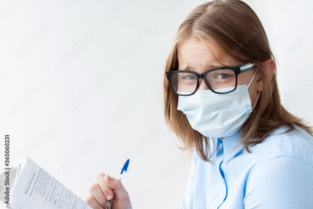 A teenage girl, blonde, in a blue shirt, a medical mask and glasses on a white background looks at the camera, holding a pen and a notebook A teenage girl, blonde, in a blue shirt, a medical mask and glasses on a white background looks at the camera, holding a pen and a notebook