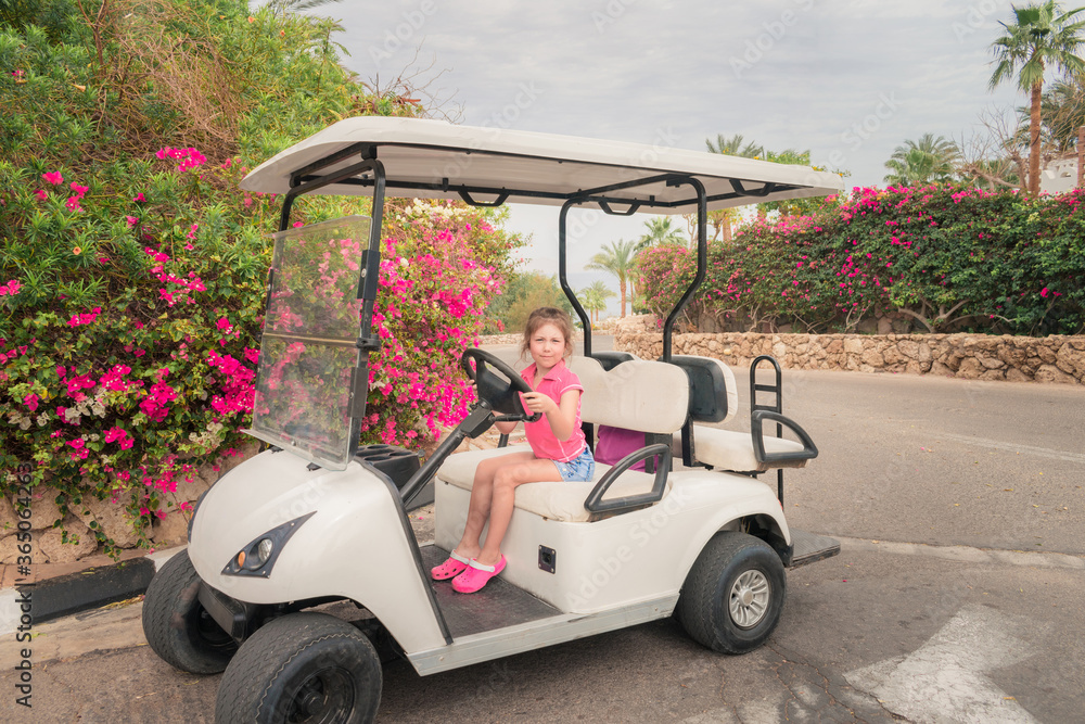 A child is playing a driver sitting on an electric car. Little girl is sitting behind the wheel of a golf cart. Little tourist has fun at a tropical resort