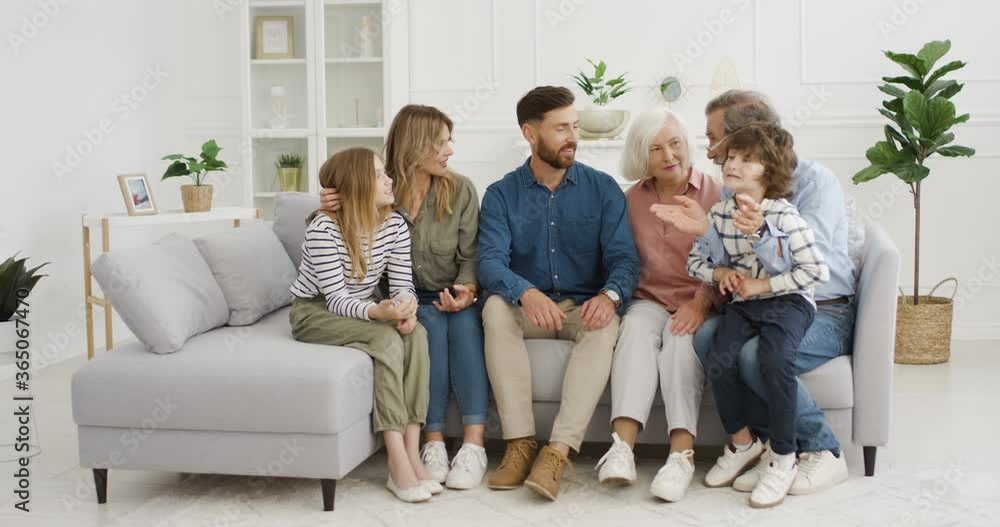 Big Caucasian family sitting on couch in living room and communicating. Grandparents, parents and small children at home on lockdown. Kids talking with mother, father, grandmother and grandfather. Big Caucasian family sitting on couch in living room and communicating. Grandparents, parents and small children at home on lockdown. Kids talking with mother, father, grandmother and grandfather.
