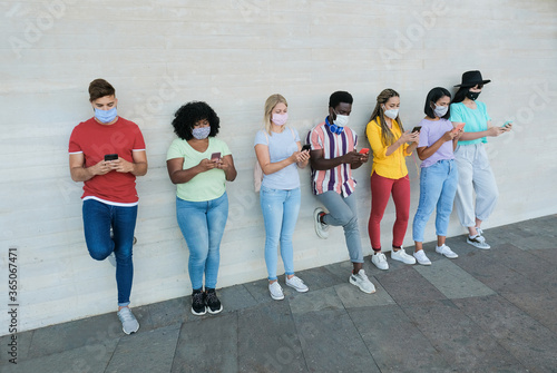 Multiracial friends standing in a raw, looking at their smartphones and wearing protective face mask - Young people having fun with technology and follow the new safety measures