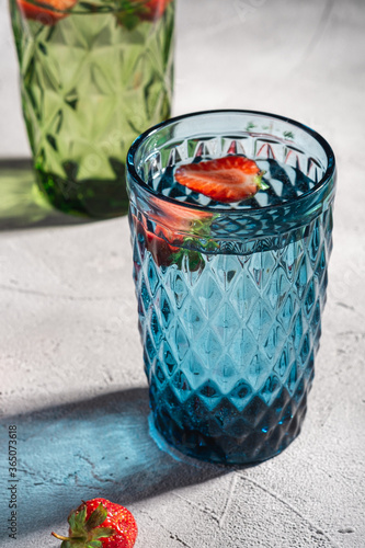 Two green and blue geometric glass cup with fresh water and strawberry fruits with colorful shadow light rays on stone concrete background, angle view