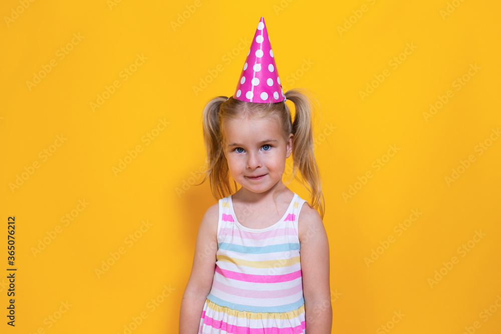 Happy birthday child girl with two ponytales in pink cap on colored yellow background.
