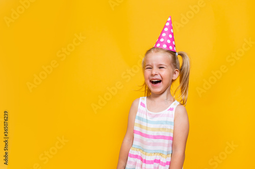 Happy birthday child girl with two ponytales in pink cap laughing on colored yellow background. Space for message.
