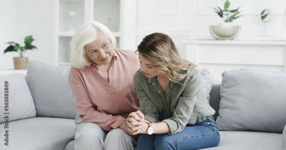 Young Caucasian woman in despair sitting on couch and worrying at home and senior gray-haired mother calming her down. Adult daughter in depression talking with old mom. Depressed desperate female. Young Caucasian woman in despair sitting on couch and worrying at home and senior gray-haired mother calming her down. Adult daughter in depression talking with old mom. Depressed desperate female.