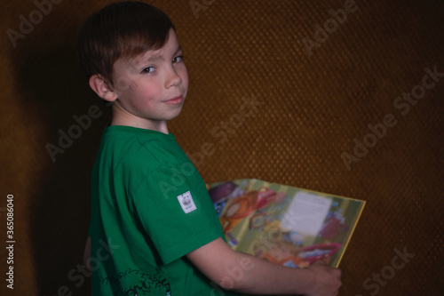 portrait of a pensive nine year old blond boy in a green t-shirt on brown background reading a yellow book with bright pictures. home schooling 