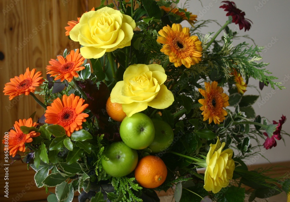 Flower arrangement with a fruit theme featuring apples, oranges, yellow roses and orange daisies