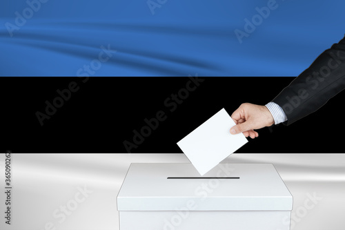 Election in Estonia. The hand of man putting his vote in the ballot box. Waved Estonia flag on background. 