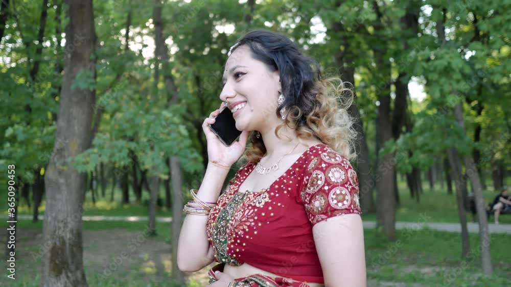 Young smiling woman in ethnic Indian red Sari talking on the phone in summer Park Young smiling woman in ethnic Indian red Sari talking on the phone in summer Park