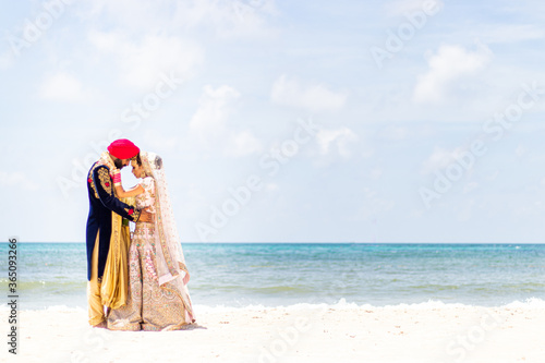Sikh bride and groom wearing bright traditional clothing on clear sandy beach beneath sunny blue skies