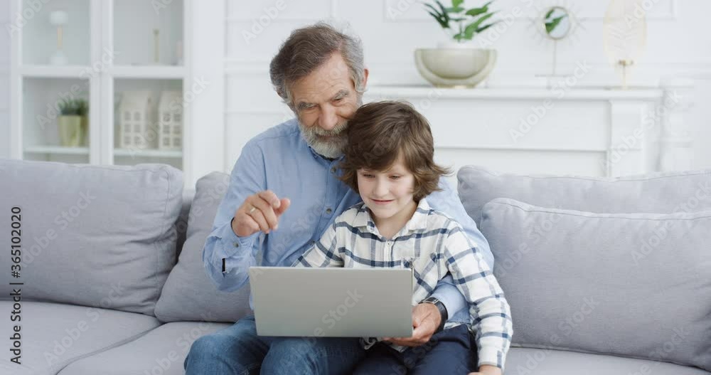 Caucasian grandfather sitting on couch in living room and teaching small cute grandson using laptop. Little boy typing on computer and playing game. Child spending day with grandpa and sitting on laps Caucasian grandfather sitting on couch in living room and teaching small cute grandson using laptop. Little boy typing on computer and playing game. Child spending day with grandpa and sitting on laps