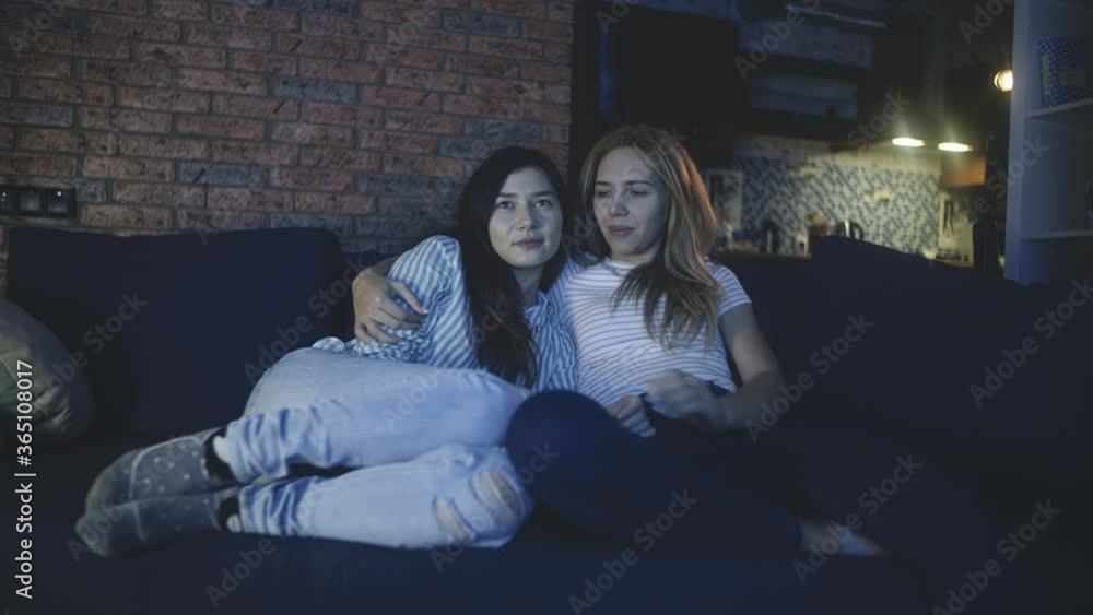 Two mixed race girls sharing love and support. Multiethnic lesbian couple lying on sofa or couch, watching movie, hugging and laughing. Concept of LGBTQI, LGBT pride month, friendship, gay family. Two mixed race girls sharing love and support. Multiethnic lesbian couple lying on sofa or couch, watching movie, hugging and laughing. Concept of LGBTQI, LGBT pride month, friendship, gay family.