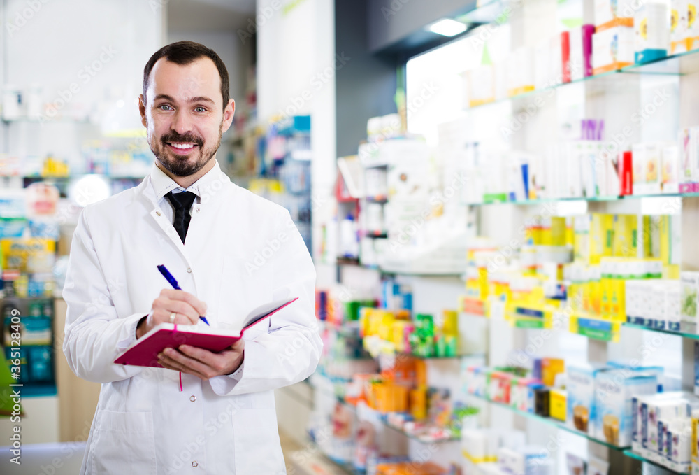 Young smiling male pharmacist noting assortment of drugs in drugstore