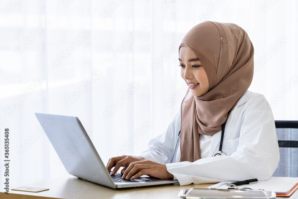 Confident Muslim female woman medical doctor sitting at desk, working typing with laptop computer in clinic hospital office Confident Muslim female woman medical doctor sitting at desk, working typing with laptop computer in clinic hospital office
