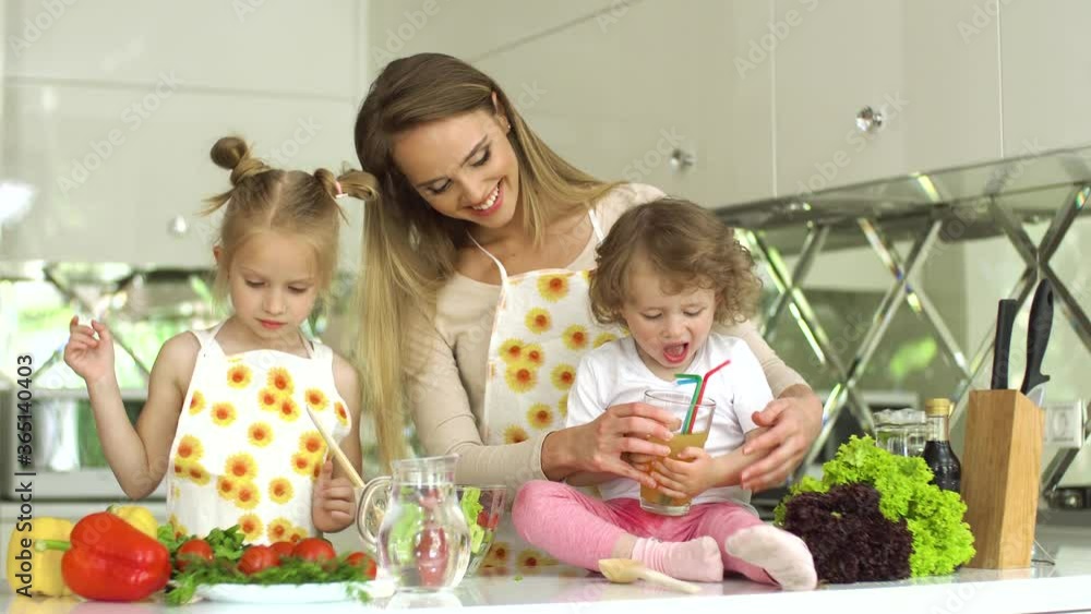 Woman With Kids Preparing Vegetable Salad At Modern Kitchen. Girl Cutting Veggies, Boy Drinking Juice From Glass Woman With Kids Preparing Vegetable Salad At Modern Kitchen. Girl Cutting Veggies, Boy Drinking Juice From Glass