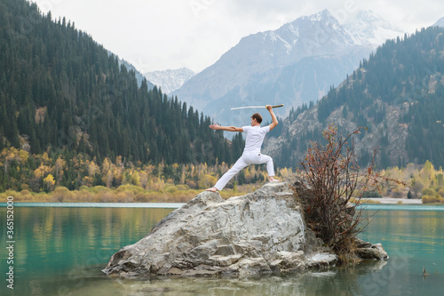 A man in white clothes with a sword stands on a large stone among the highlands.