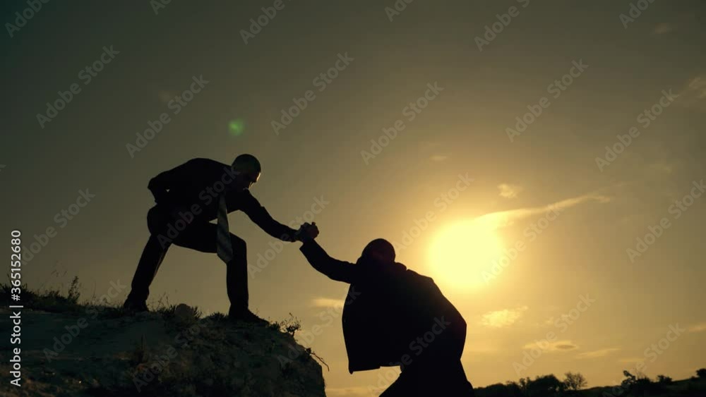 Businessmen climb to the top of the mountain, careers and one of them gives a helping hand. Silhouette of two men at sunset. Dressers help in crisis, overcoming obstacles. The concept of teamwork.