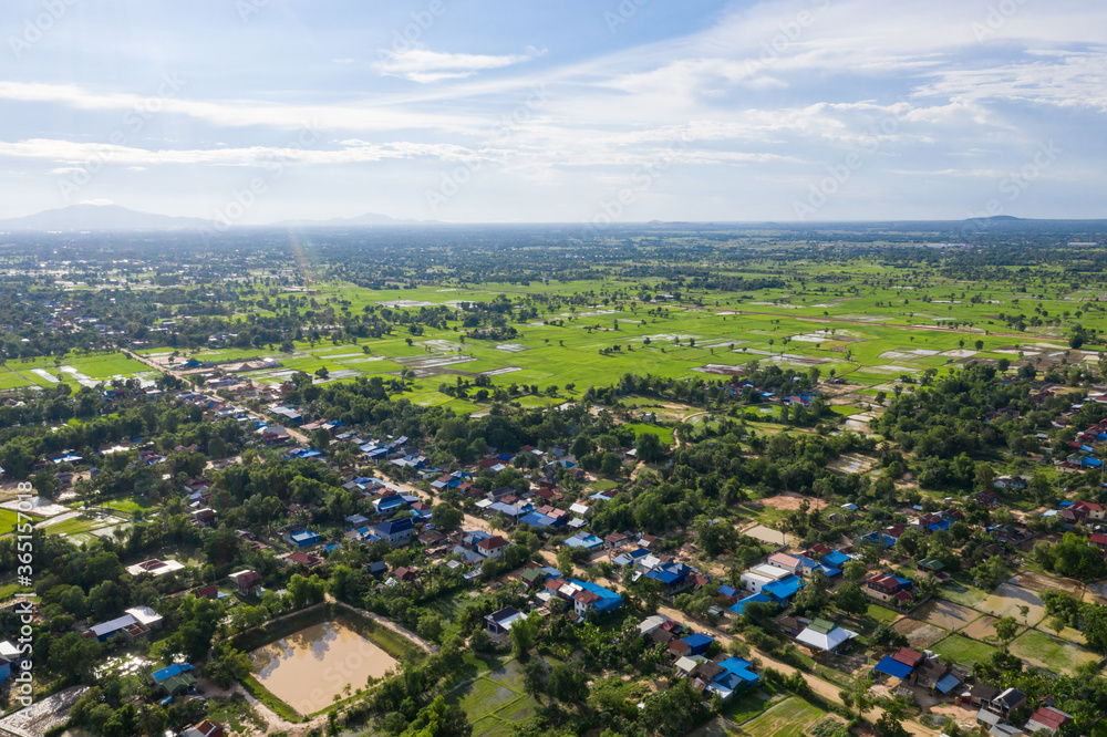 A top down aerial view of a small country town with traditional houses with orange roofs, a red dirt road, rice fields, and palm trees in the jungle in Cambodia.