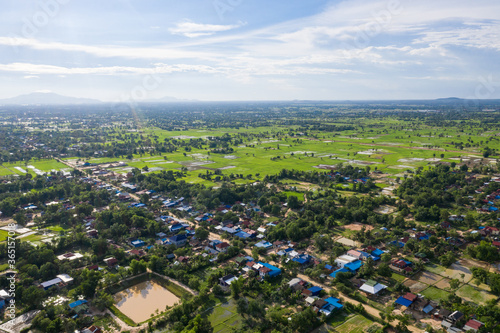 Fototapeta Naklejka Na Ścianę i Meble -  A top down aerial view of a small country town with traditional houses with orange roofs, a red dirt road, rice fields, and palm trees in the jungle in Cambodia.
