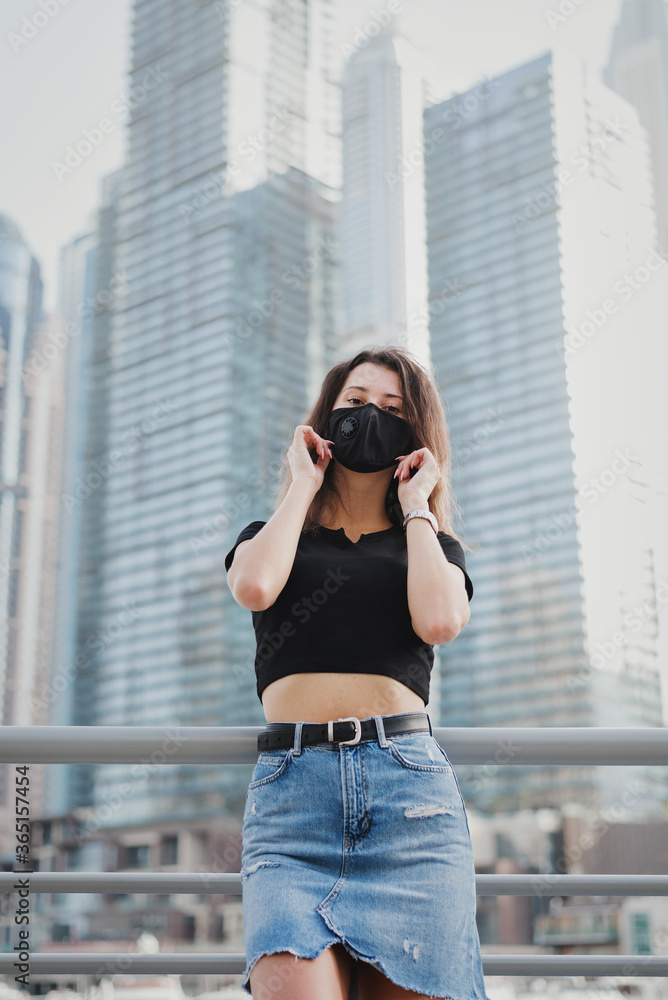 Young European woman wearing protective face mask with filter and standing outside near city center with office buildings and skyscrapers. New normal during Covid 19 pandemic