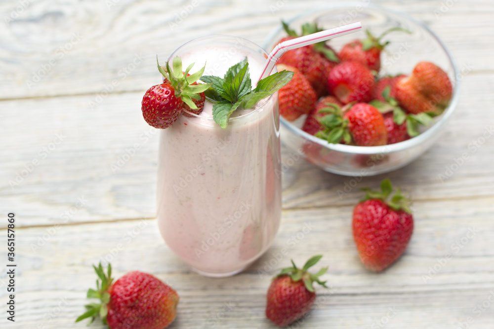 A glass glass with a strawberry milkshake with a straw, stands on a light wooden background, the berries are in a transparent vase and on the table A glass glass with a strawberry milkshake with a straw, stands on a light wooden background, the berries are in a transparent vase and on the table