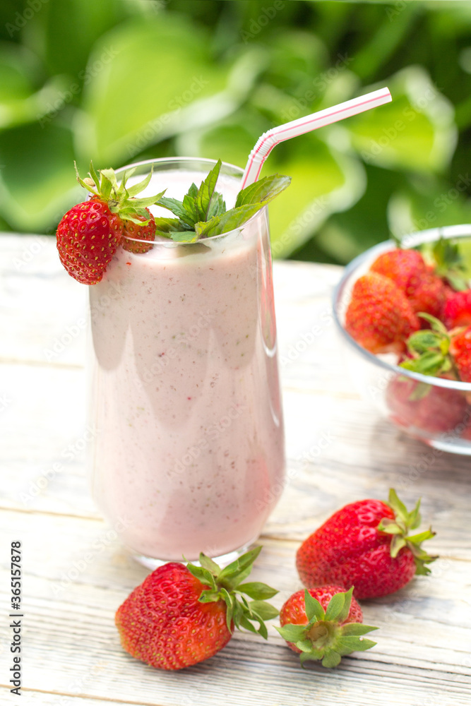 A milkshake of fresh strawberries stands on a light wooden background , strawberry berries are on the table and in a transparent vase A milkshake of fresh strawberries stands on a light wooden background , strawberry berries are on the table and in a transparent vase