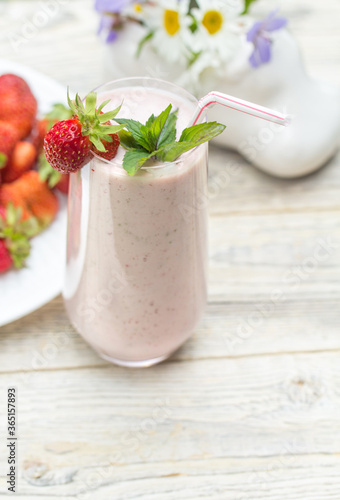 A glass glass with a strawberry milkshake and a white plate with strawberries stand on a light wooden background