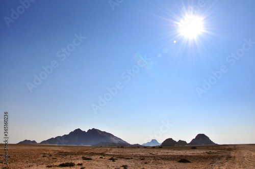 Minimalist landscape of harsh hot desert with Spitzkoppe Mountains on the horizon  vivid blue no clouds sky and sun shining  Namibia  Africa