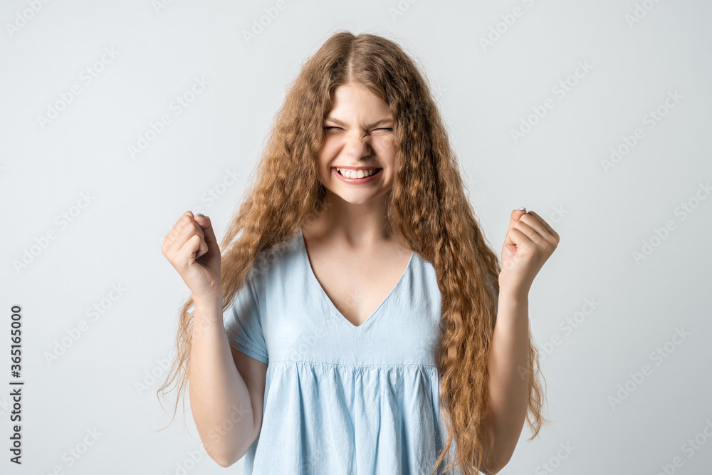 YES! Life is wonderful! Joyful girl with curly long hair keeps fists clenched and exclaims in triumph, celebrates success, isolated over white background YES! Life is wonderful! Joyful girl with curly long hair keeps fists clenched and exclaims in triumph, celebrates success, isolated over white background